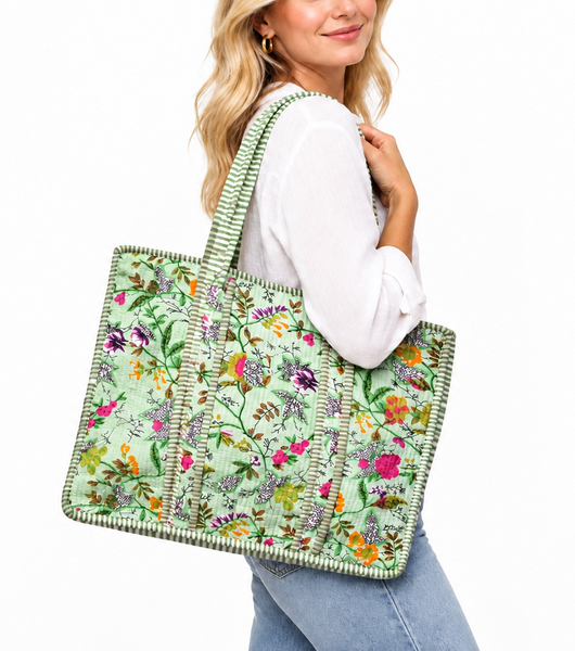 A woman wearing a white long-sleeve top and light blue jeans carries a large woven tote bag with a light green floral pattern and striped green-and-white handles, photographed against a plain white background.