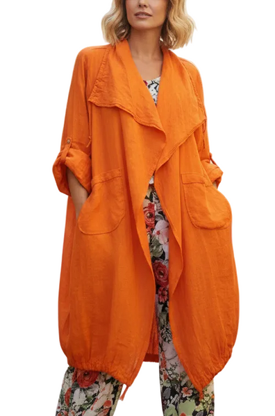 A woman modeling a bright orange oversized linen-blend duster coat with large patch pockets and draped sleeves, paired with floral pants, against a plain white background.