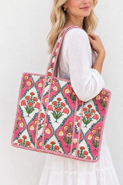 A woman wearing a white dress carries a large pink and white patterned tote bag with floral motifs and green accents against a plain white background.