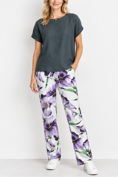 A woman modeling a dark gray short-sleeve top and white pants with a purple floral print, standing against a plain white studio background.