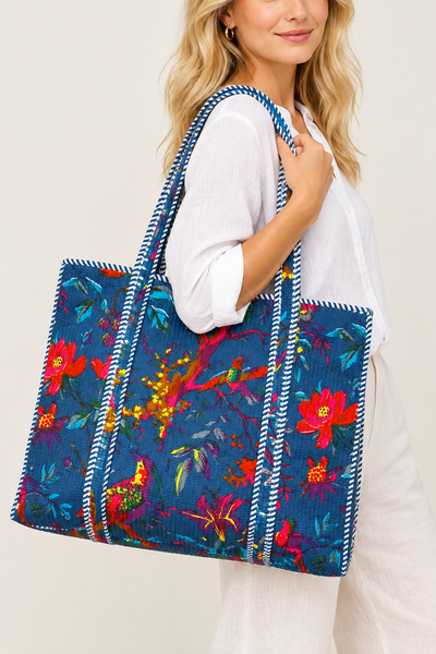 A woman wearing a white long-sleeve top and white pants carries a large blue tote bag decorated with a colorful floral and bird print, set against a plain light beige background.