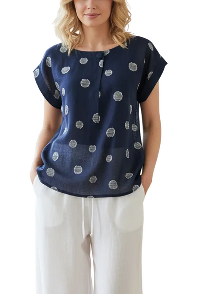 A woman stands in a bright, sunlit room with wood floors and white walls, wearing a navy blue short-sleeve blouse with white circular motifs and white wide-leg pants.