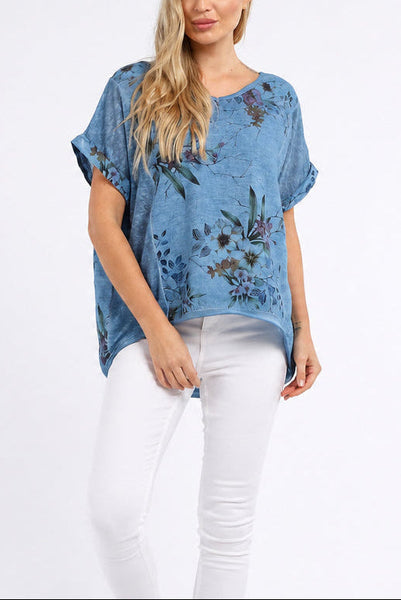 A woman modeling a short-sleeve blue top with a floral print, paired with white skinny jeans and white sneakers, against a plain white studio background.