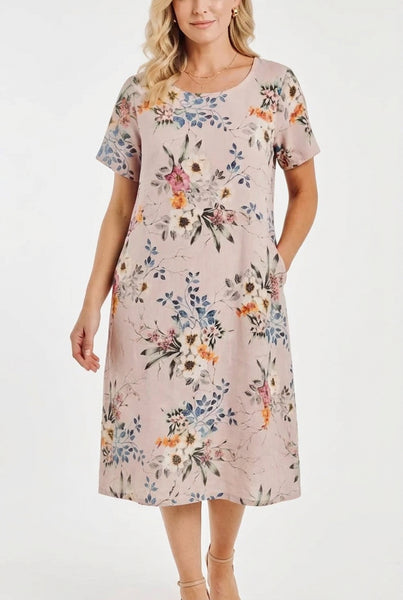 A woman modeling a short-sleeve knee-length shift dress with a delicate multicolor floral print on a dusty pink background, paired with tan flat sandals, against a light gray studio backdrop.