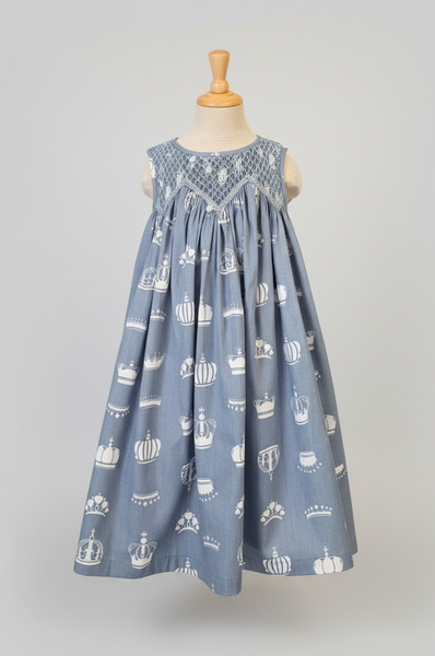 A sleeveless baby dress on a mannequin, featuring a dusty blue fabric with a white repeating pumpkin and crown pattern, and a smocked bodice.