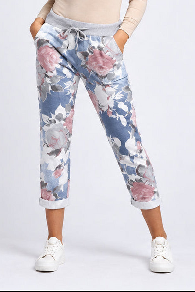 A person wearing floral jogger-style pants with a blue base, pink and white flowers, and a gray drawstring waistband, paired with white sneakers against a light gray studio background.