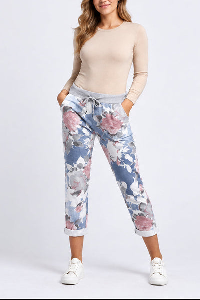 A woman modeling a beige long-sleeve top and blue floral cropped sweatpants with white sneakers, standing against a light gray studio background.